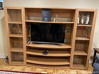 Full frontal view of a modular wooden wall unit with two glass door cabinets on the sides and open shelves in the center holding a flat-screen TV and a few small items on shelves.