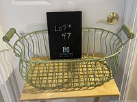 Wire basket painted pale green with wooden handles on both ends, photographed from front and side angle in front of a door.