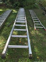 Full length view of the 24-foot aluminum extension ladder laid out on grass next to two other ladders and lawn equipment.