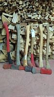 Six axes in front of stacked lumber, showing variations in handle shape, length, and axe head design and color.