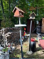 Outdoor image showing two bird feeders on poles with bases and a black and red painted milk can next to garden gnome decorations and firewood stacks.