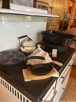 Various cast iron cookware items including mini skillets with wooden bases, frying pan, griddle with circular molds, floral tea pot, egg pan, spatula, and roasting pans displayed on a stove.