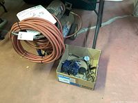 The air compressor on floor with attached orange coiled hose and instruction manual on top, next to cardboard box with metallic accessories.
