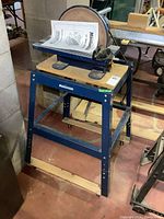 Side view of Mastercraft 12-inch disc sander mounted on its blue metal wheeled stand with wooden base, showing overall structure and manual on top.