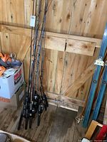 Wide shot of 17 fishing poles with grips and reels clustered together, leaning against wooden wall.