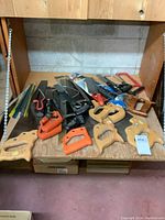 Wide view of multiple hand saws with various handles (wooden and plastic), laid on wooden shelf, some saw frames and spare blades visible