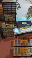 Photo of multiple stacked storage containers and large multi-drawer units holding assorted small hardware parts including nails, screws, nuts, and bolts. Items placed on floor with basement background.