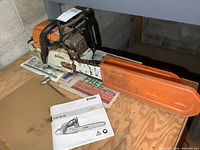 Side view of chainsaw on wooden surface with orange bar guard installed and instruction manual