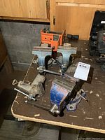 Four metal vises arranged on a wooden workbench, showing two identical smaller blue vises, one larger blue bench vise, and one unpainted clamp-style vise.