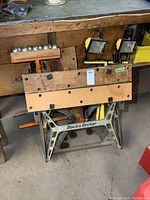 Front view of Black & Decker folding Workmate with wooden clamp surfaces, roller stand, and two yellow work lights in background