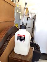 White translucent plastic 5.5 gallon utility jug with black cap and metal pouring spout placed on a wooden bench in a garage environment.