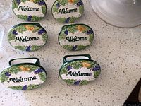 Six ceramic napkin rings arranged on a white speckled countertop, each oval ring with "Welcome" text and painted flowers and grapes, green handle on each.