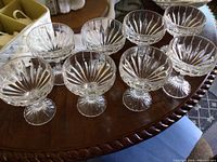 Eight clear crystal drinkware glasses arranged on a round wooden table, each with a faceted cut design and footed base.