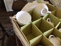 Eight white coffee cups arranged in a protective box with green padded dividers. One cup is placed on top, displaying the embossed floral pattern and handle.
