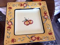 Top view of square ceramic serving platter showing red peaches in center and decorative floral and fruit border in gold, red, and green tones.
