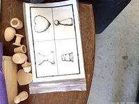 Photo showing a set of wooden 3D shapes next to a laminated card with black-and-white outline drawings of the shapes for identification.