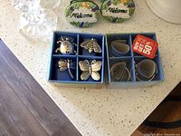 Photo showing two blue boxes on a countertop each containing four silver plated napkin rings. One box holds insect-shaped napkin rings (ladybug, bee, dragonfly, butterfly) and the other holds four plain round napkin rings. Additional items such as small decorative signs and a glass vase are seen partly in the photo.