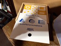 Wooden game board showing raised tactile yellow shapes and numbers with an instruction sheet labeled 'Touch 'N Play Lotto Shapes & Numbers' by ZipDeck.