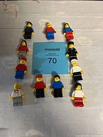 Top view of eleven 1980s LEGO minifigures arranged around a blue MaxSold card with lot number 70. Figures mostly wear red, blue, black, or white outfits with some having hats or caps.