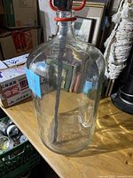 Large clear glass jug with red handle, clamp-style stopper visible on top, approx 22 inches tall