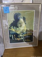Framed art print of a girl sewing leaning against a wall indoors, showing the full artwork and frame with protective wrapping.