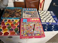 Five assorted jigsaw puzzles displayed on a surface, showing varied box artwork including Bingo, colorful shaved ice, ceramic pots, kaleidoscope pattern, and crossword puzzle themes.