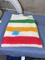 Image of folded wool blanket, showing white base and red, yellow, and green stripes with a blue lot tag on top.