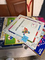 Stack of six loose game boards shown from an angle with Anti-Monopoly board on top, colors and spaces visible on several boards.