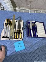 Two cutlery sets displayed on a blue surface, one steak cutter set with black and yellow handles in a gold-tone case, another child's set in blue case with white-handled knife.