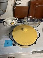 Photo of a white ceramic gravy boat with floral decorations, a clear glass pie plate, and yellow ceramic covered serving dish in black metal holder.
