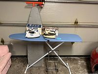 Photo showing two ironing tables, one with gray padded cover and the other with blue padded cover, with three irons placed on top and a metal ironing cart in front.