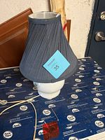 White ceramic table lamp with black pleated fabric shade shown unlit with a blue lot tag marked 35 on lampshade.