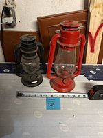 Two oil lanterns side by side on a flat surface with measuring tape showing about 10 inches width total. One lantern is red marked with 'Dietz', the other is black with no visible brand.