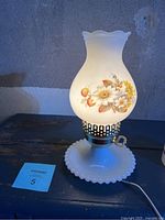 Vintage milk glass electric table lamp illuminated showing floral painted glass shade and scalloped edges on shade and base.