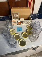 Full view of six used clear plastic Oster Mini-Blend containers each with a gold-colored lid and three new containers in original box packaging plus instruction booklet.