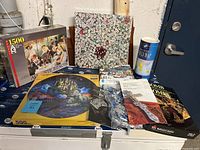 Lot of six assorted jigsaw puzzles stacked on a surface, including a 1500-piece Auguste Renoir art puzzle, a 500-piece circular castle puzzle, a dice-themed puzzle, and a cylindrical container puzzle.