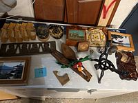 Overview of mixed decorative arts and crafts including framed pictures, wooden and metal sculptures, small carved objects, and a wooden fan.