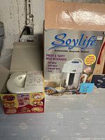 Front view of Soylife Automatic Soymilk Maker box and the white appliance beside it showing lid and spout area, along with rice cooker box below.