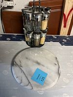 Photo shows a black metal spice rack with 12 glass jars filled with spices and silver lids, placed on a table next to a round white marble lazy Susan.