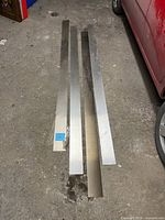 Three stainless steel kick plates laid on a concrete floor next to a red vehicle, showing weathered, dull finish and straight edges.