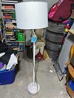 Full view of one white floor lamp with white cylindrical shade, standing on a round base, in a cluttered room with board games and tires in background.