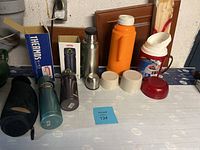 Photo showing six assorted thermoses and insulated jugs on a table, includes a black quilted carrying case thermos, teal green metal thermos, purple Thermos brand vacuum bottle in original box, large orange insulated jug, red and white jug, small silver cup/flask.
