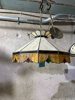 Side angle view showing the stained glass panels in cream and amber colors with scalloped lower edge in amber with red and green diamond-shaped accents. Hanging hardware and chain suspension visible.