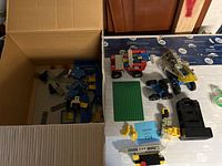 Box of vintage Lego pieces including base plates, loose bricks, minifigure parts and wheels, and partially assembled space vehicles on a white surface.
