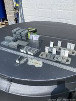 Various galvanized steel deck building materials arranged on black surface under natural light including joist hangers, angle brackets, hurricane ties, and a box of coated screws.