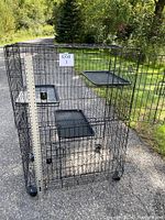 Tall black metal 3-tier indoor cat cage playpen with plastic floor pans on each tier, one large door open, located outdoors on pavement near grass.