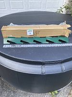 Four green powder-coated steel compost bin brackets laid out with a cardboard box behind them on a black round surface, showing packaging and lot label.