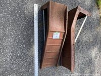 Two rusted metal car ramps side by side on asphalt with a measuring tape for size reference.