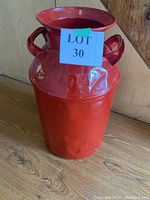 Full view of the vintage red painted metal milk can showing side handles and open top.