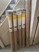 Five rolls of PVC window blinds standing upright near staircase. Four labeled 48x72 inches and one larger 72x72 inches blind. Beige wood-like style slats visible through plastic.